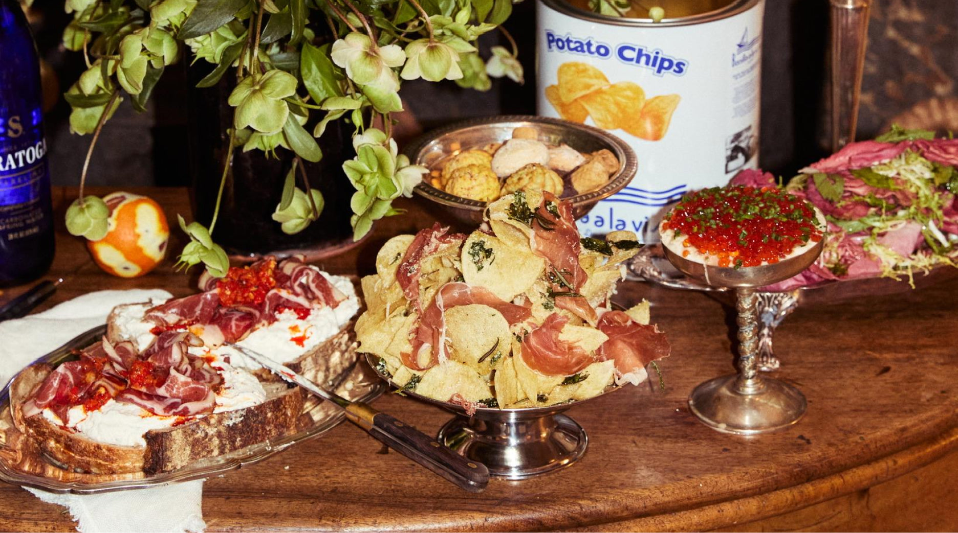 antique silver bowls filled with potato chips and slices of prosciutto and amaretto cookies next to a platter of thick bread topped with fresh mozzarella cheese and salame