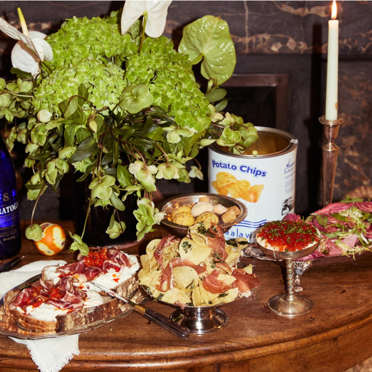 antique silver bowls filled with potato chips and slices of prosciutto and amaretto cookies next to a platter of thick bread topped with fresh mozzarella cheese and salame