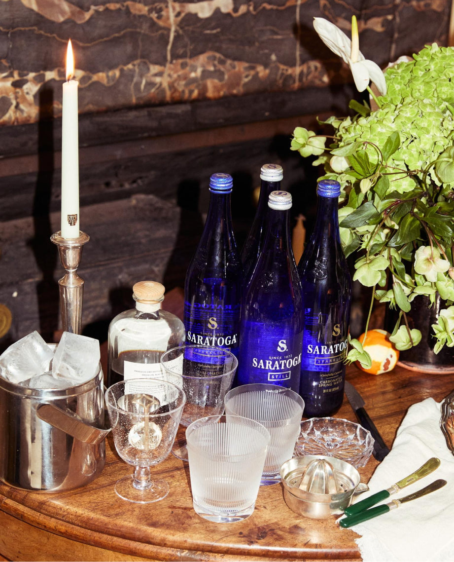 wood side table with cocktail station including bottles of saratoga spring water and mixers and garnishes next to a silver ice bucket and a thin tapered candle