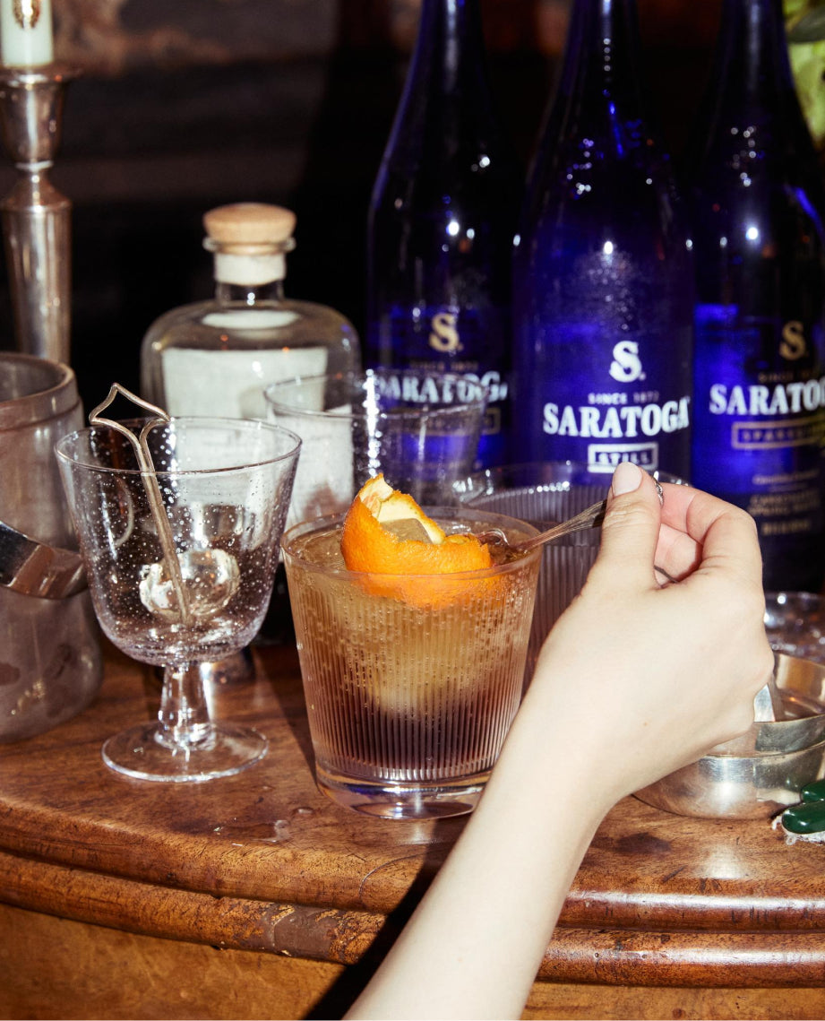 bottles of amaro and saratoga sparkling water next to a hand stirring a cocktail with an orange garnish